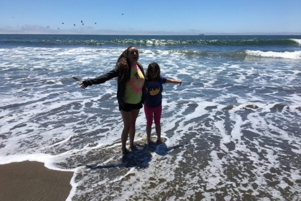 Maegan and Sara standing nexnt to each other in the surf on the beach with arms outstretched. Maegan is holding a feather in her hand.